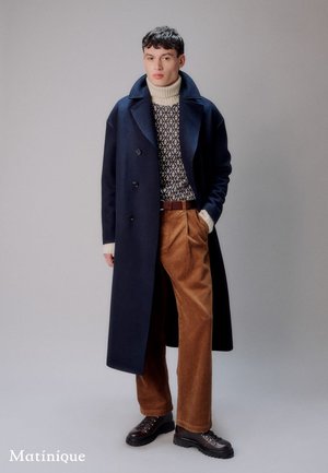 Navy overcoat with large lapels and buttons, beige knit sweater, tan corduroy pants, and black lace-up shoes stand against a light background.