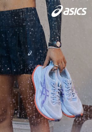 Baskets de course violettes claires avec un design texturé, fermeture à lacets et accents orange, tenues par une personne en tenue de sport devant un arrière-plan humide.