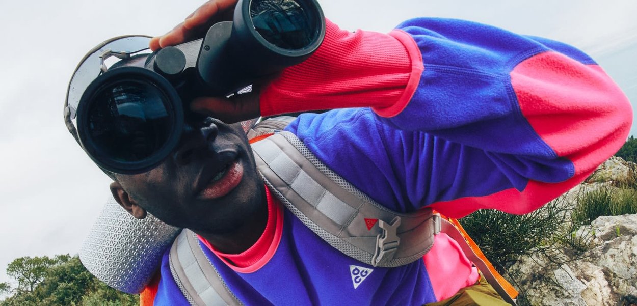Persona con chaqueta morada y rosa y mochila, mirando a través de binoculares al aire libre en un terreno rocoso.