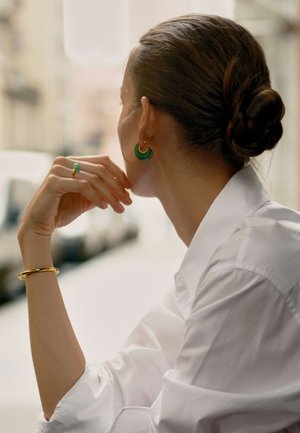 Boucles d'oreilles en vert et or, chemise blanche, bracelet en or et bague en pierre verte. Cheveux coiffés en un chignon bas, main positionnée de manière réfléchie.