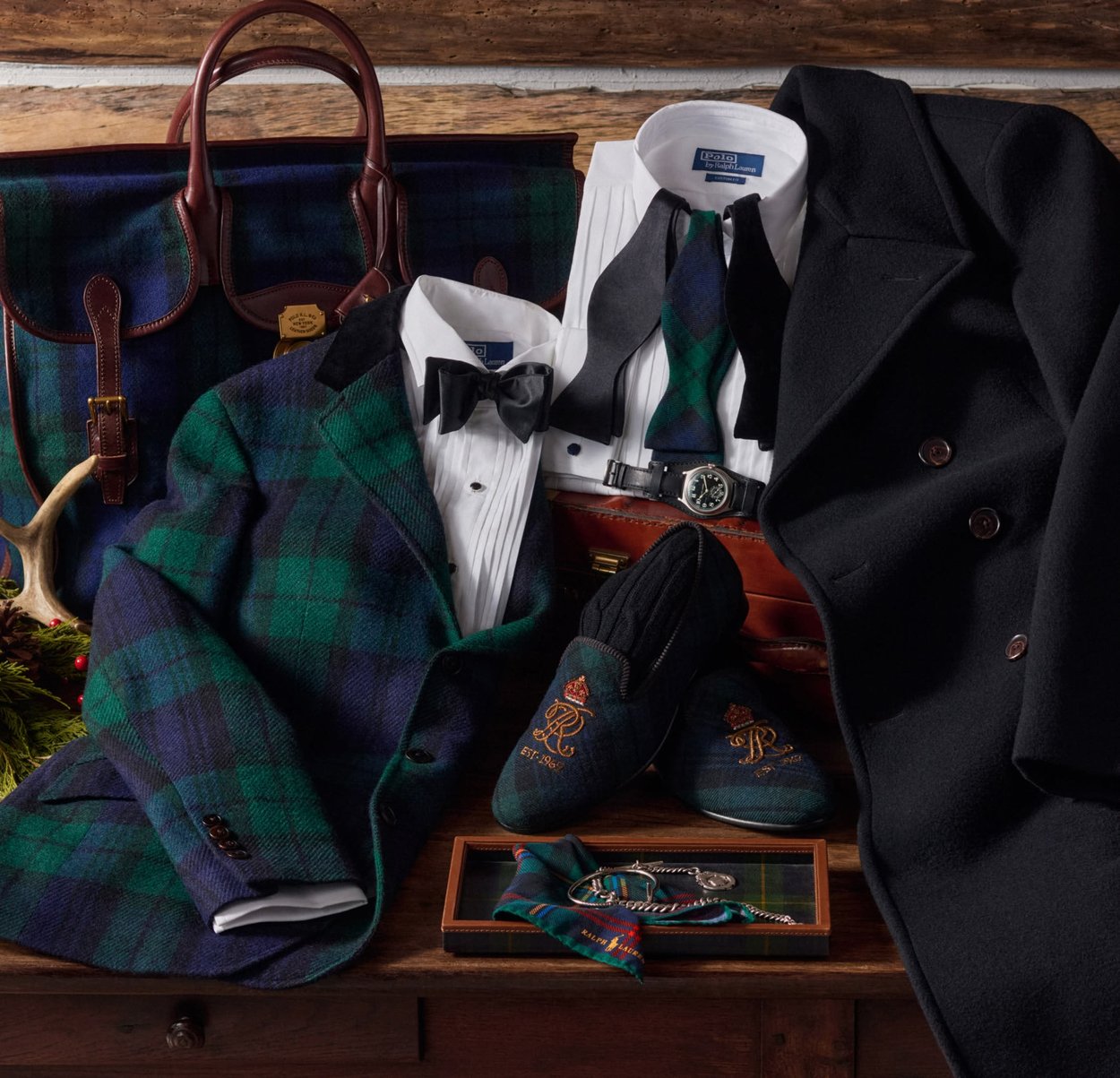 Vihreä- ja laivastonsiniruudullinen tweed-takki, valkoiset paidat, rusetti, musta takki, nahkalaukku, monogrammilla varustetut tohvelit ja asusteet esillä puisella pöydällä.