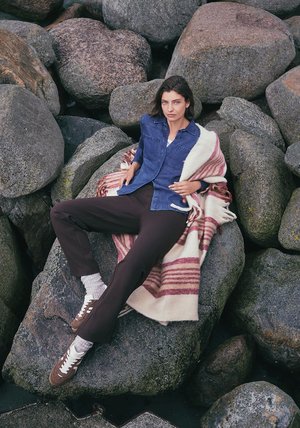 Denim overhemd over een wit topje, donkerbruine broek en grijze sokken. Gewikkeld in een crèmekleurige en roze gestreepte deken, zittend op gestructureerde stenen.