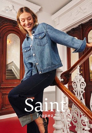 Frau lächelt und lehnt sich auf ein hölzernes Geländer, trägt eine Jeansjacke und weit umgeschlagene Jeans, in einem dekorierten Innenraum mit Holztüren.