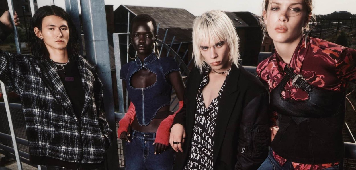 Quatre modèles posent devant une structure métallique. Les tenues incluent une veste à carreaux, un haut en denim avec découpes, une chemise à motifs floraux, et un vêtement noir ajusté avec des éléments en métal.