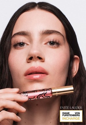 Giovane donna con labbra rosa lucide tiene vicino al viso un tubetto di gloss rosa con tappo dorato su sfondo neutro.