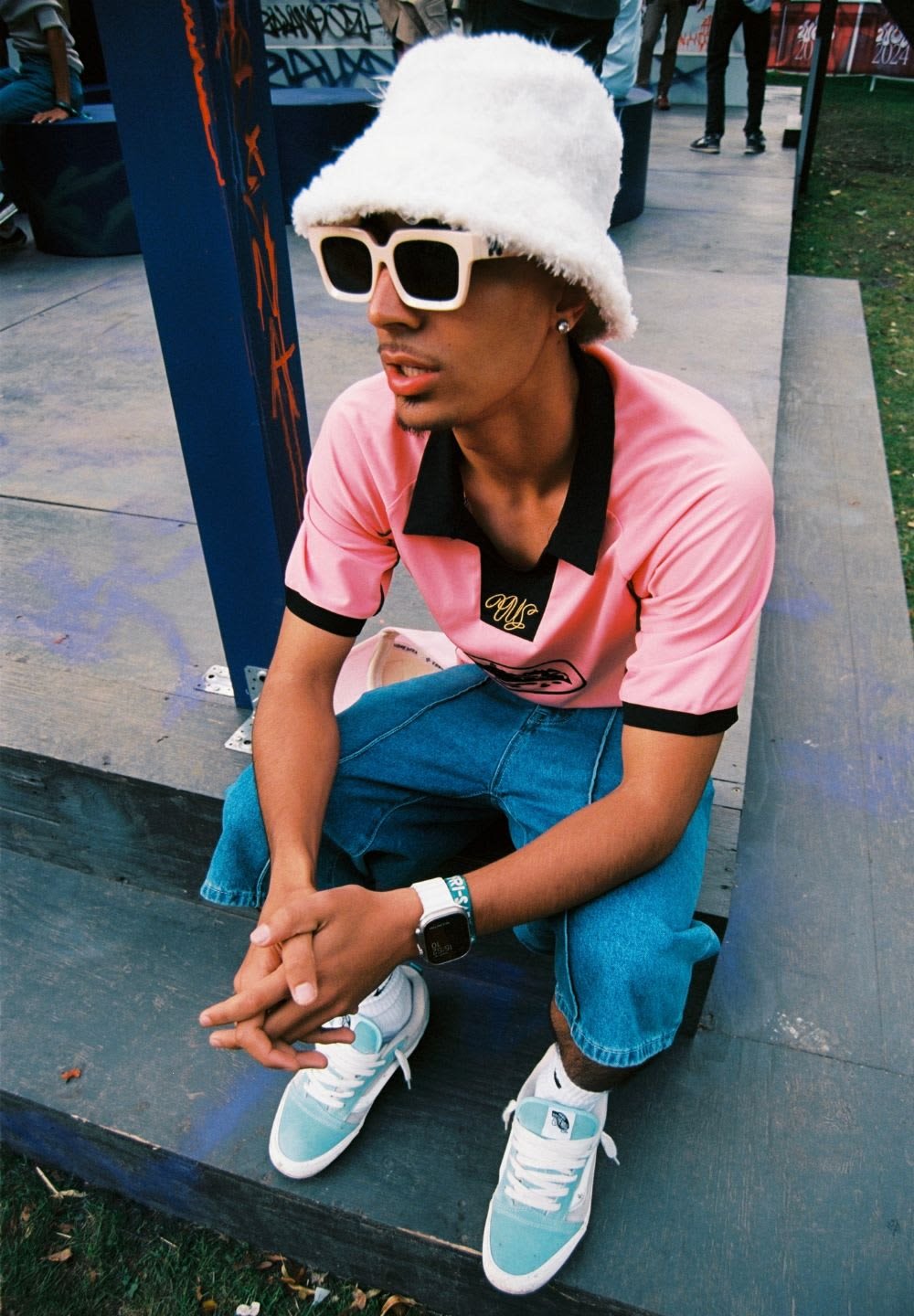 Rosa polo-t-shirt med svarta detaljer, blå jeans, vit bucket hat och vita/gröna sneakers. Person sitter på en träyta, med händerna korslagda.