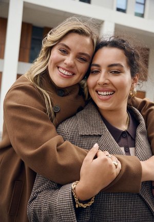 Deux femmes sourient l'une à côté de l'autre. L'une porte un manteau marron à boutons ; l'autre un blazer à motif pied-de-poule sur une chemise sombre.