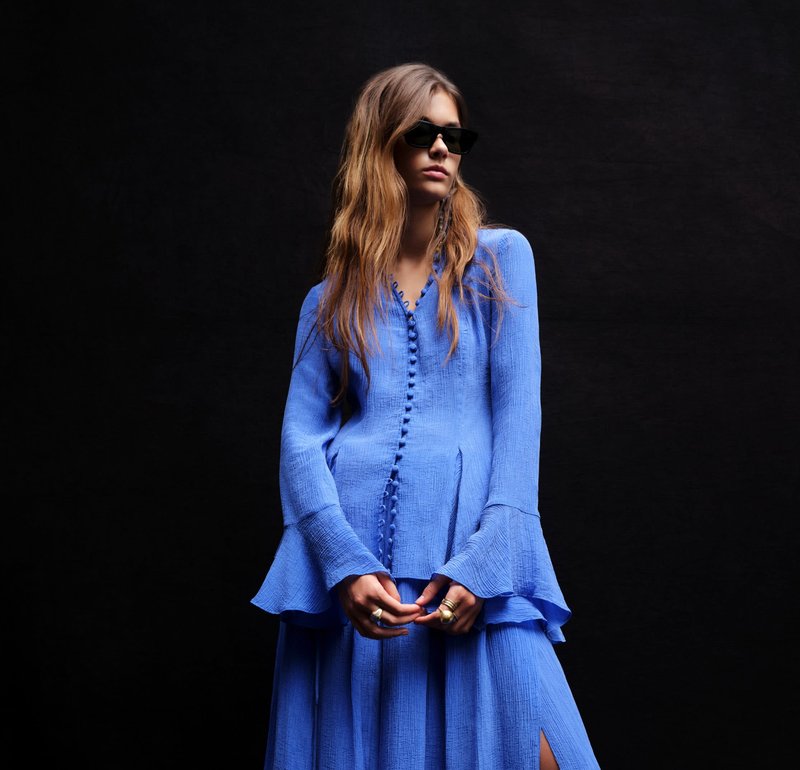 Vrouw met lang haar die zwarte zonnebril draagt en een gestructureerde blauwe jurk met knopen en wijde mouwen, tegen een zwarte achtergrond.