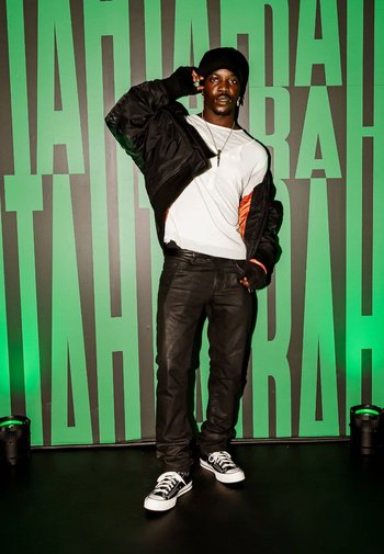 Black bomber jacket with red accents over a white t-shirt, black jeans, and black and white sneakers. Background features large green text.