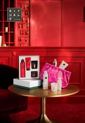 Sett med skjønnhetsprodukter i en gaveeske. Inneholder røde og svarte beholdere, en rosa pose, hvite lys og et rundt trebord med gullaksenter.