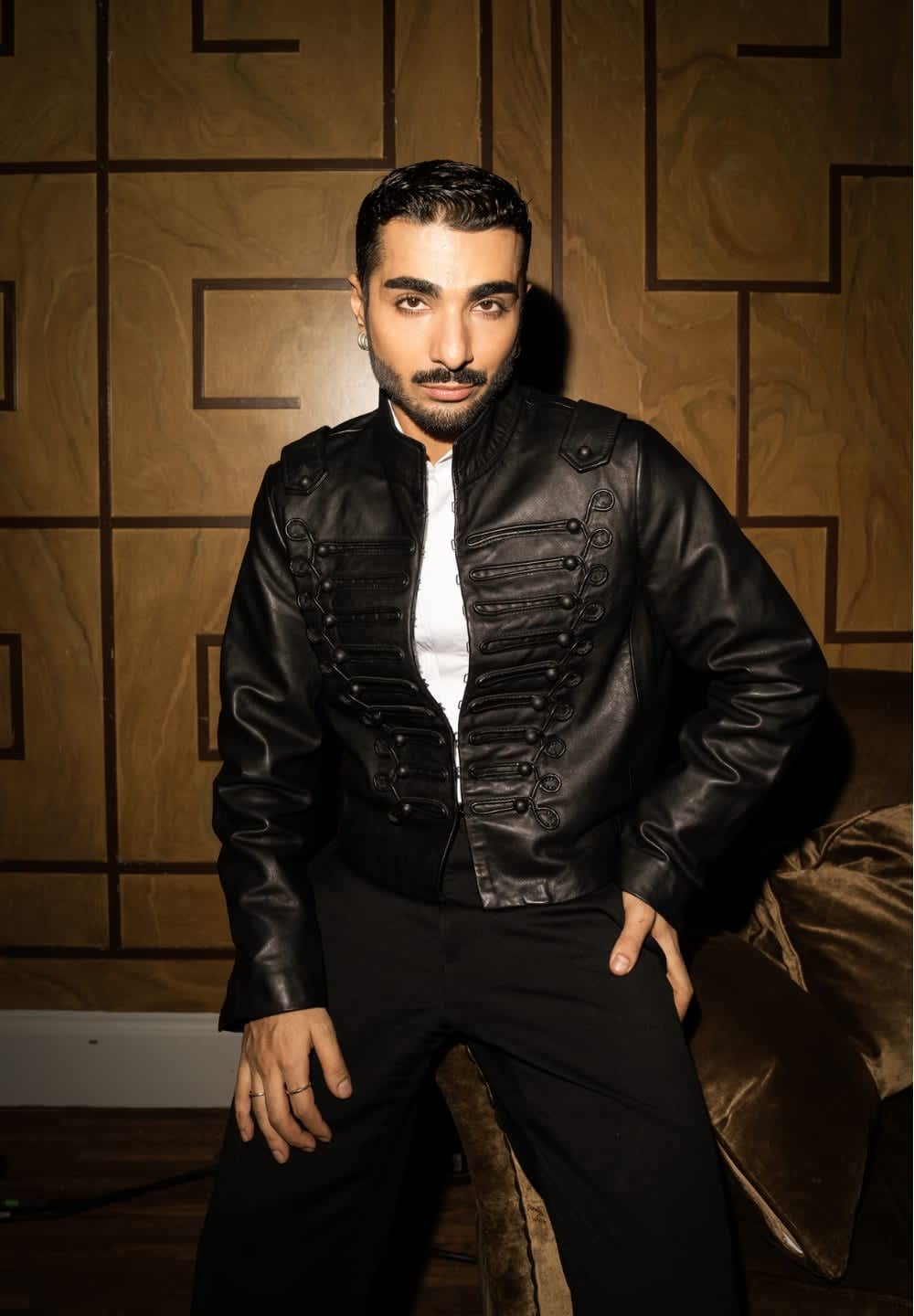 Svart lädercroppad jacka med militärinspirerade knappar, bärs över en vit skjorta, kombinerad med svarta byxor; texturerad bakgrund.