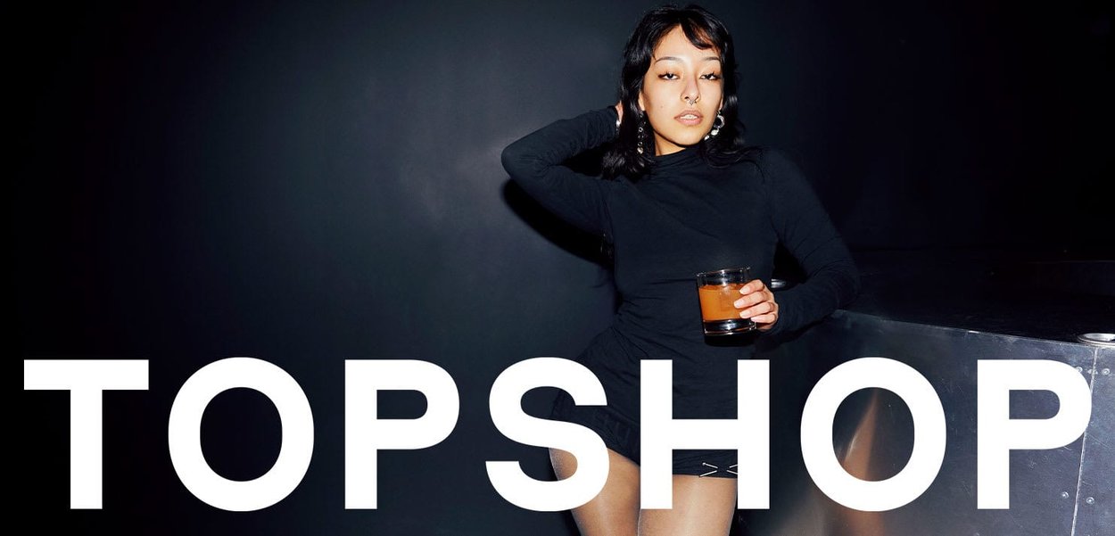 Woman in black turtleneck and shorts holding a drink, posing confidently against a dark background with "TOPSHOP" text below.
