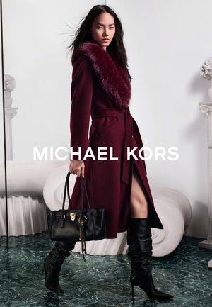 Frau trägt einen bordeauxroten Mantel mit Pelzkragen, schwarze lässige kniehohe Stiefel, hält eine schwarze Lederhandtasche und steht auf einem grünen Marmorboden.