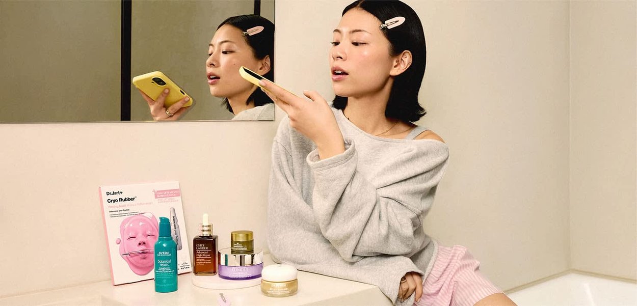 Donna con felpa grigia e fermaglio per capelli che tiene un telefono giallo, seduta accanto al bancone del bagno con prodotti per la cura della pelle accanto al riflesso nello specchio.