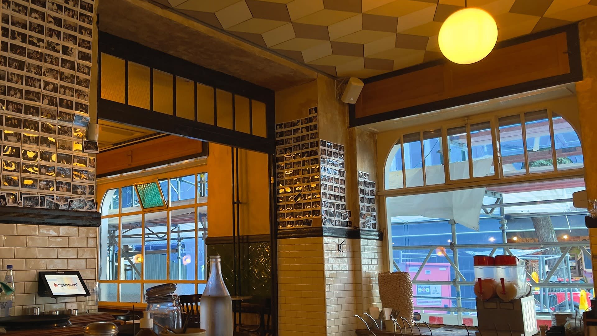 Interieur van een café met betegelde muren, een hexagonaal patroon in het plafond, ingelijste foto's, grote ramen en een ronde hanglamp.