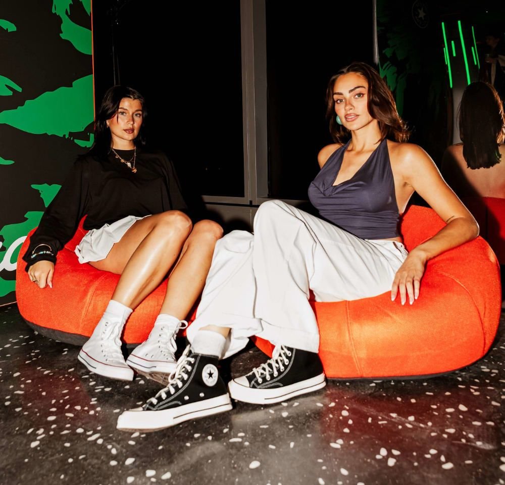 Two individuals sit on a bright orange bean bag chair. One wears a black long-sleeve top and a white skirt; the other, a navy halter top and white pants.