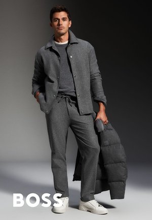 Man in een grijze jas, bijpassende broek en witte sneakers houdt een grijze pufferjas vast tegen een donkere gradient achtergrond.