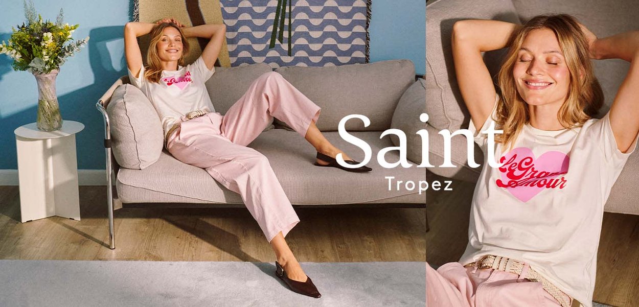 Frau trägt weißes grafisches T-Shirt und rosa Hose, lehnt sich auf einem grauen Sofa in einem hellen Raum zurück, neben ihr steht eine Vase mit Blumen.