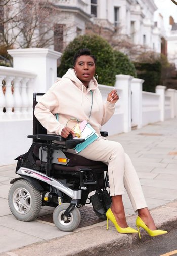 En elektrisk rullstol med silver- och svarta accenter, utrustad med tjocka hjul. Användaren har på sig en ljus huva och beige byxor och håller en färgglad väska.