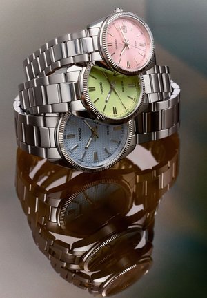 Tres relojes de pulsera Casio apilados con correas metálicas plateadas y esferas redondas en tonos pastel rosa, verde y azul sobre una superficie reflectante.
