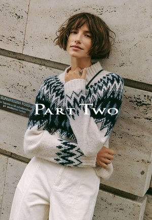 Junge Frau mit kurzen braunen Haaren, die einen gemusterten Wollpullover und weiße Hose trägt, lehnt draußen an einer Steinmauer.