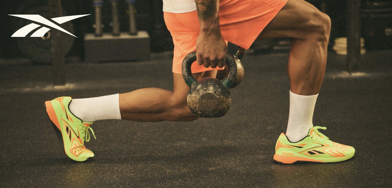 Persoon in oranje korte broek tilt een kettlebell met beide handen, draagt neon geelgroene sneakers en witte sokken op de gymvloer.