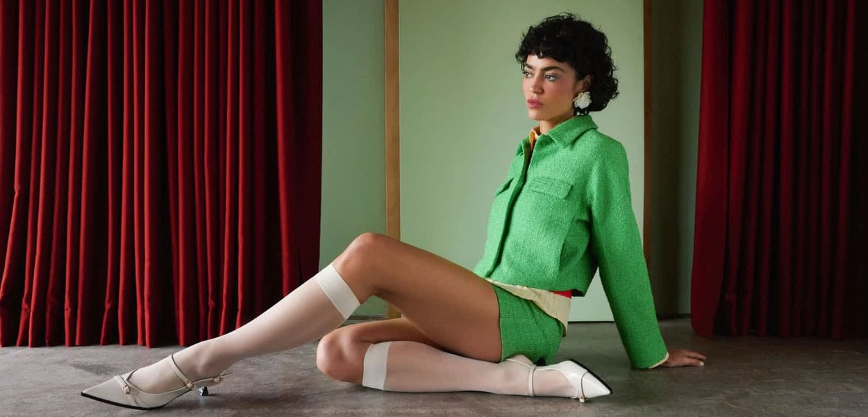 Veste et shorts texturés verts, bas blancs montants jusqu'à la cuisse, et escarpins blancs pointus avec détails en métal. Rideaux rouges en arrière-plan.