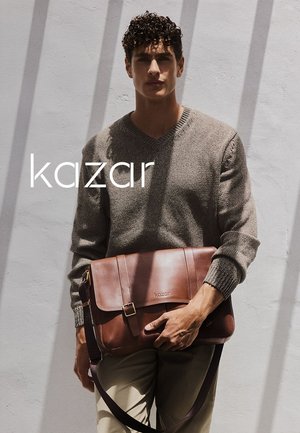Braune Leder-Umhängetasche mit Klappe, verstellbarem Tragegurt und eingeprägtem Logo. Getragen von einem Mann in einem grauen Pullover vor einem hellen Hintergrund.