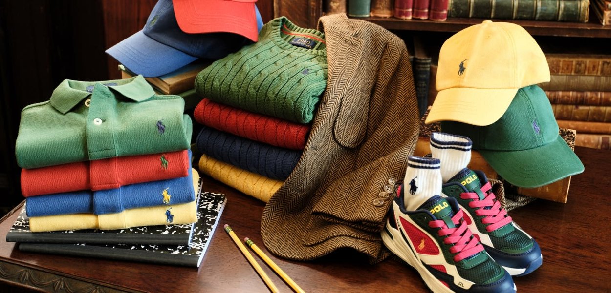 Gefaltete bunte Poloshirts, gemusterter Pullover, braune Jacke, drei Baseballkappen, ein Paar Turnschuhe mit Socken, Bleistifte und Notizbücher auf einem Holztisch.