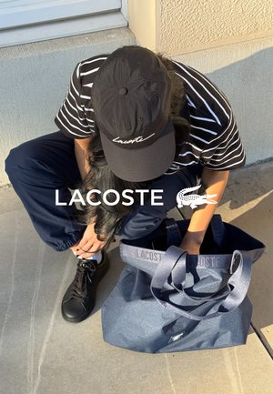 Marine totebag met Lacoste-branding, stoffen handvatten en een gestructureerd oppervlak. Zwarte pet met wit logo en gestreept shirt. Zwarte sneakers.
