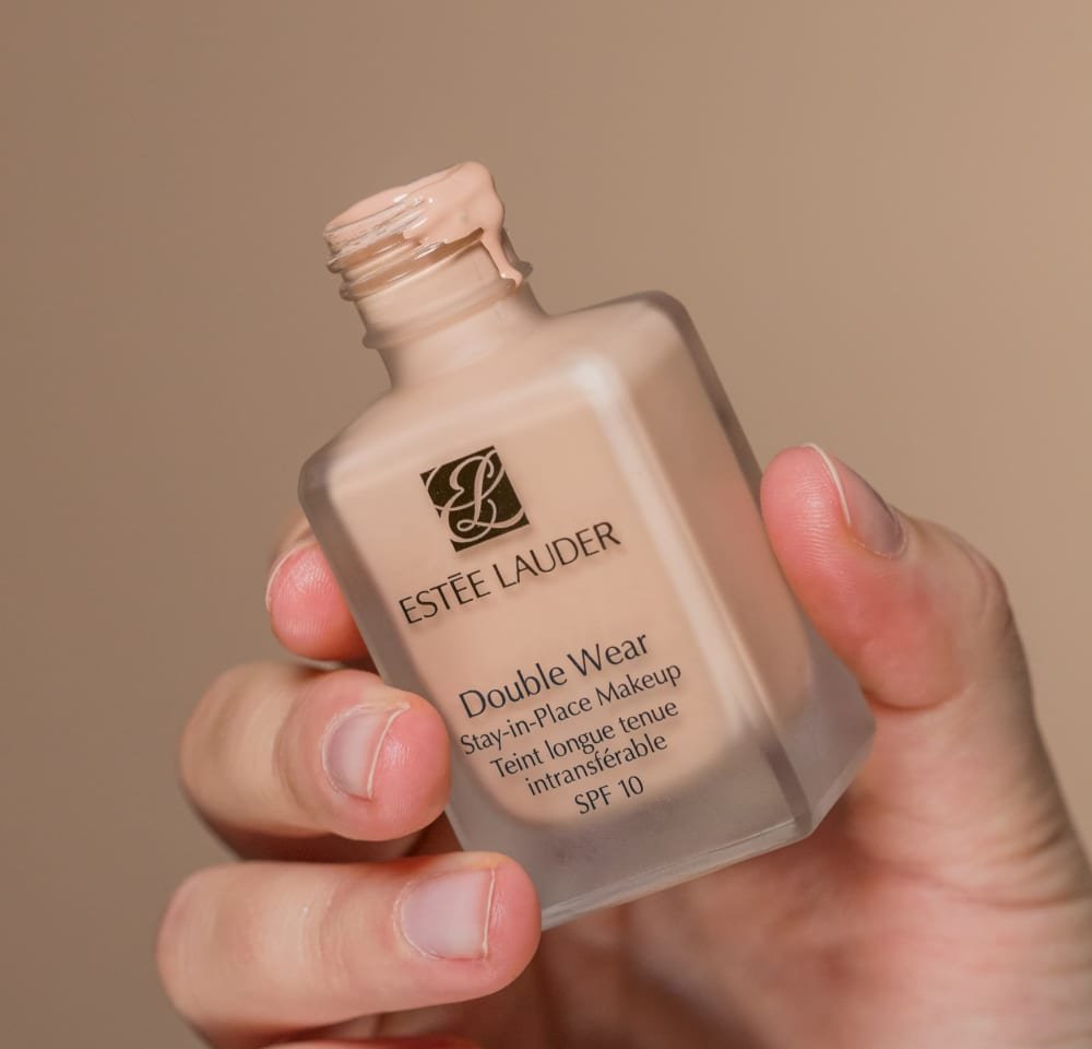 Estee Lauder Double Wear foundation i en klar glasflaske, beige væsketekstur, med et sort logo, holdt i en hånd. SPF 10 angivet.