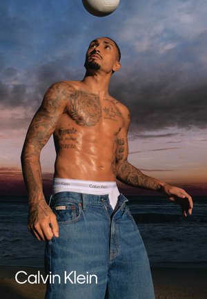 Skjorteløs tatoveret mand i løse jeans og Calvin Klein undertøj headers en fodbold ved solnedgang på en strand.