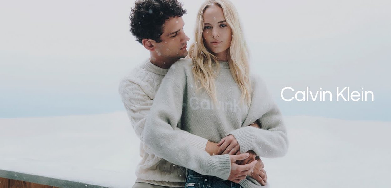 Dos modelos se abrazan al aire libre con ropa de invierno. La mujer lleva un suéter gris claro y el hombre un suéter de punto trenzado color crema.