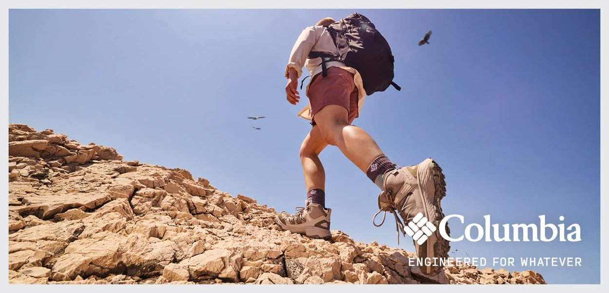 Wanderschuhe in hellem Beige, mit texturierter Sohle für Grip, getragen mit beigen Shorts und einem weißen Langarmshirt, während er ein felsiges Terrain unter klarem Himmel erklimmt.
