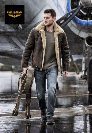 Veste bombardier en cuir marron avec un col en fourrure, pull gris, jeans bleus et bottes noires. Tient une bandoulière dans la main gauche, pavé mouillé.