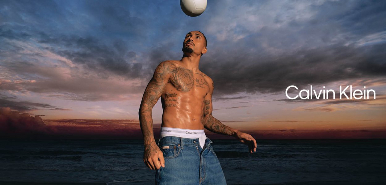 Uomo senza maglietta con tatuaggi, indossa biancheria intima Calvin Klein e jeans, mantiene in equilibrio un pallone da calcio contro un cielo al tramonto sul mare.