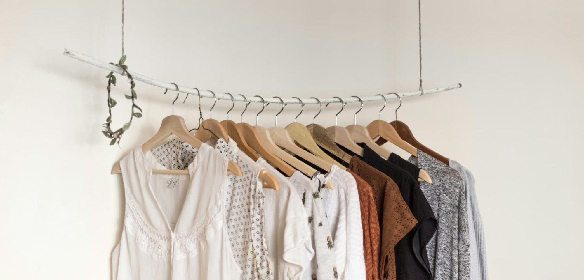 Nine garments suspended on wooden hangers: assorted colours (white, beige, brown, black, grey), textures, patterns, and sleeve styles on a curved rod.