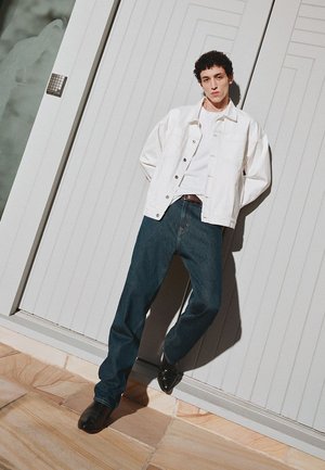 Junger Mann lehnt an einer weißen Täfelungstür und trägt eine weiße Jacke, ein weißes Hemd, dunkelblaue Jeans und schwarze Schuhe.