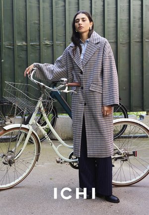 Cappotto lungo a scacchi con colletto oversize, motivo blu e bianco, abbinato a pantaloni a righe. Sullo sfondo è presente una bicicletta.