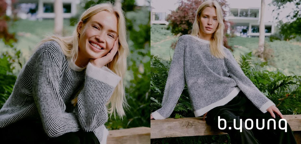 Grau gestreifter Pullover mit weißem Saum, bietet eine lässige Passform und strukturiertes Material. Modell posiert im Freien zwischen Grünanlagen. Markenname sichtbar.