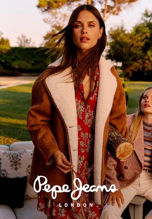 Donna con giacca di montone marrone e abito rosso a fiori tiene un tronco all'esterno, con un'altra donna seduta vicino che guarda verso l'alto.