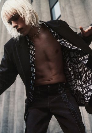 Blazer negro sobre una camisa estampada con texto; jeans negros; modelo con cabello rubio platino. Enfócate en el diseño gráfico distintivo de la camisa.