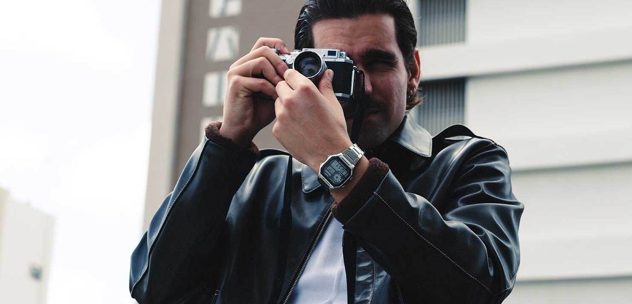 Veste en cuir noir, chemise blanche, portant une montre numérique. Tenant un appareil photo vintage, se concentrant sur l'objectif. L'arrière-plan montre un bâtiment.