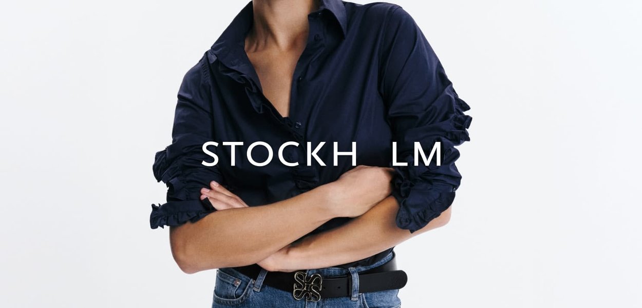 Persoon met een marineblauw geplooid overhemd met lange mouwen, armen gekruist, blauwe spijkerbroek en een zwarte riem met een decoratieve gesp.