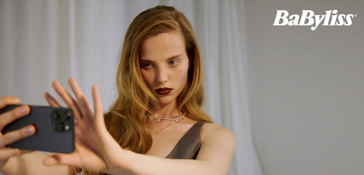 BaByliss hårstylingsverktøy på en grå overflate. En person med langt, bølgete hår tar en selfie, iført en grå topp og perlekjede.