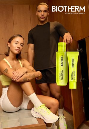 Frau sitzt in Sportkleidung und Mann steht neben ihr in einem Holzraum, mit zwei leuchtend gelben Biotherm-Hautpflegeflaschen, die neben ihnen schweben.