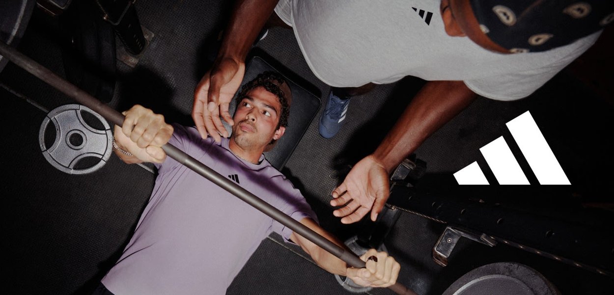 Barre avec des poids dans une salle de sport. Une personne soulève la barre tandis qu'une autre apporte un soutien par-dessus. Les deux portent des vêtements de sport avec un logo.