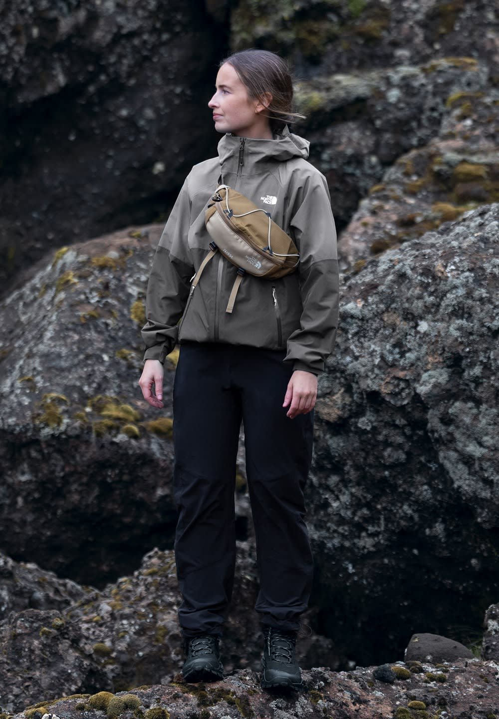 Giacca impermeabile verde oliva con dettagli grigi, pantaloni neri e scarponi da trekking neri. Una borsa a tracolla marrone aggiunge un tocco funzionale. Rocce sullo sfondo.