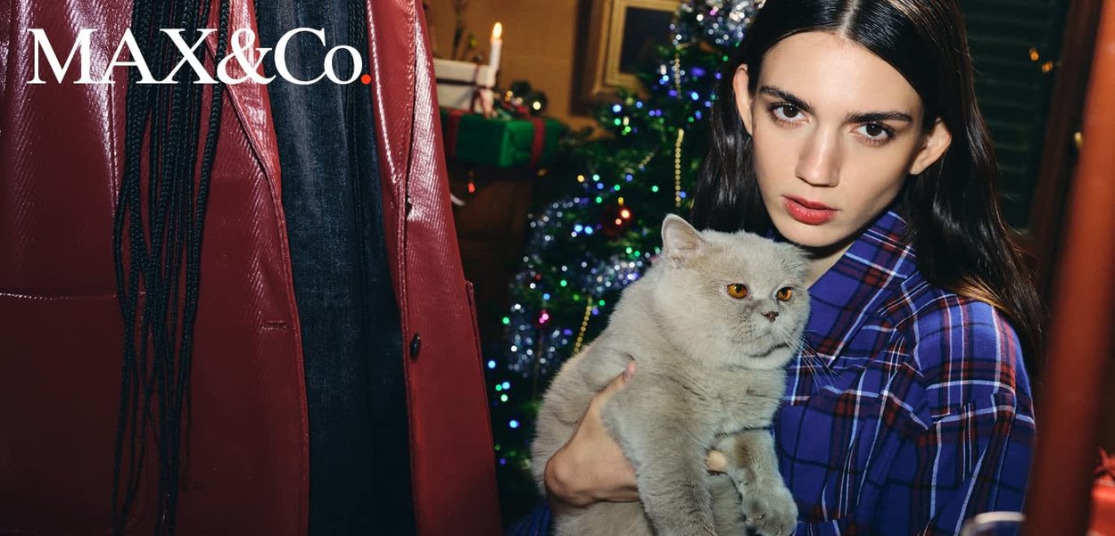 Jeune femme portant une veste à carreaux bleue tenant un chat gris et pelucheux, avec un sapin de Noël décoré et des cadeaux emballés en arrière-plan.