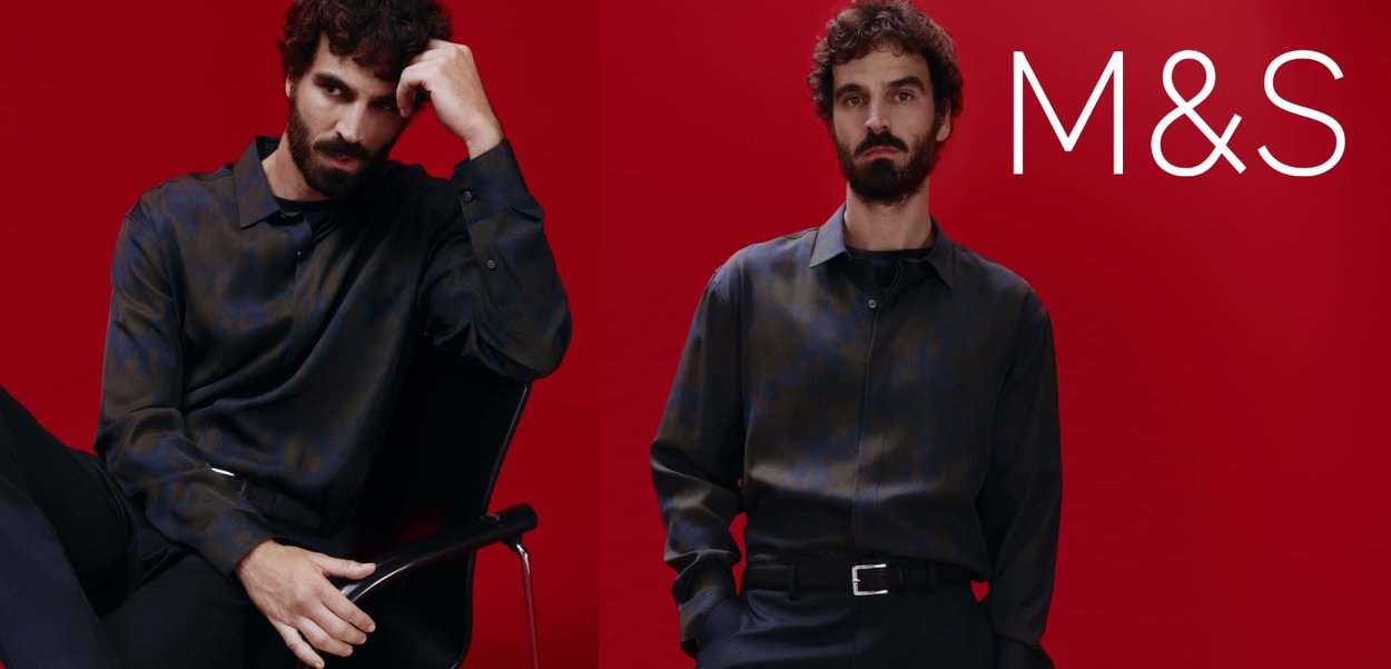 Ciemna koszula w długim rękawie z wzorem, z kołnierzykiem zapinanym na guziki, połączona z czarnymi spodniami. Tło jest jaskrawo czerwone, co podkreśla strój.